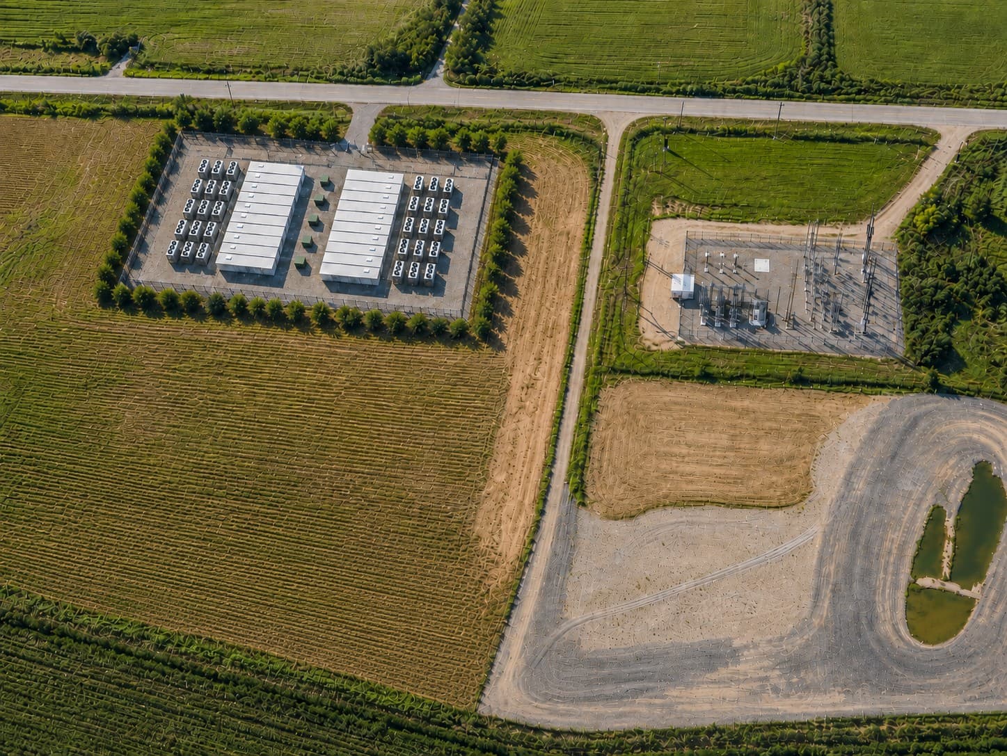 Conceptual aerial rendering of the proposed Oakwood site: two rows of low modular compute buildings flanked by cooling units, with electrical equipment between them on a fenced gravel pad, ringed by planted screening on all four sides, beside the existing electrical substation and surrounded by farm fields